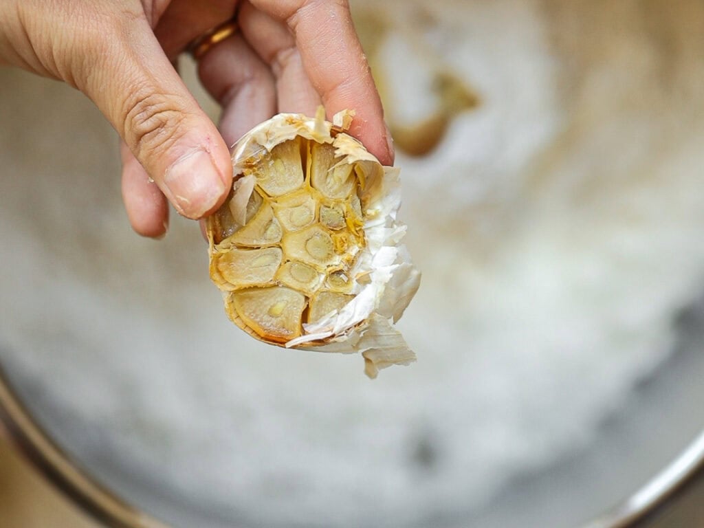 A hand holds a roasted garlic bulb with golden-brown cloves visible. The background is blurred, highlighting the garlic in the foreground.